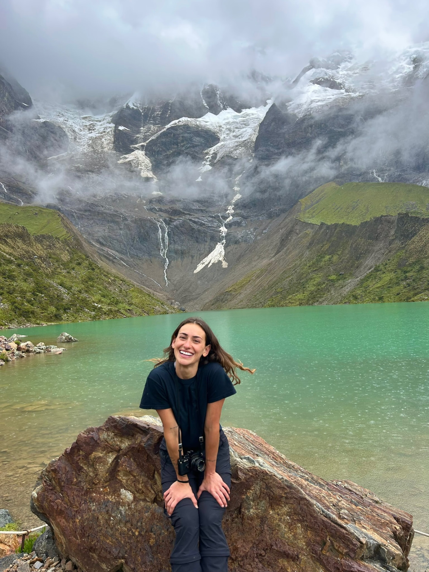 Lucia sitting on a rock beside a still mountain lake, laughing